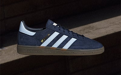 A pair of navy blue and white Adidas sneakers sits on a dark surface, with a shadowy background.