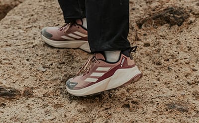 The image shows a person's feet wearing a pair of pink and white sneakers standing on a sandy, rocky surface.