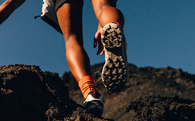 The image shows a person's legs and feet wearing hiking boots with a patterned design, standing on a rocky, mountainous terrain against a clear blue sky.