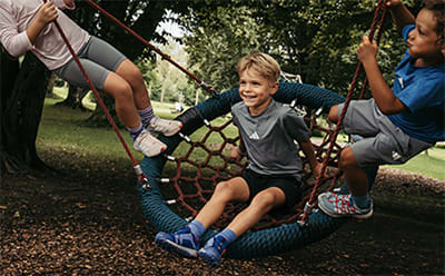 A group of children playing on a colorful net swing in a lush, green outdoor setting.