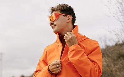 A person wearing a bright orange jacket stands outdoors against a blurred background.