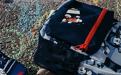 A black backpack with various patches and designs, including a red strap, sits on a colorful, textured surface in the foreground, with a blurred, natural background.