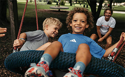 Two young children, a boy and a girl, are sitting on a swing set in a park-like setting, with trees and other people visible in the background.