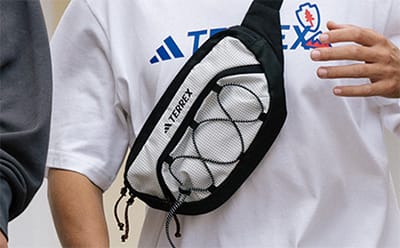 A person wearing a white sports jersey with the Adidas logo and holding a blue and white card in their hand, with a black fanny pack or waist bag visible in the foreground.