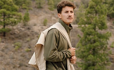 A young man wearing a beige jacket stands in a forested area, with trees and greenery visible in the background.