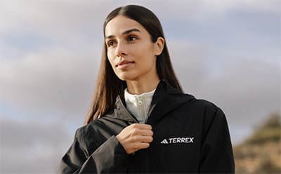 A young woman with long dark hair wearing a black Terrex jacket stands outdoors against a cloudy sky background.