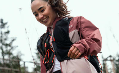 A young woman in a pink jacket and black pants stands smiling in a wooded outdoor setting, with trees and a fence visible in the background.