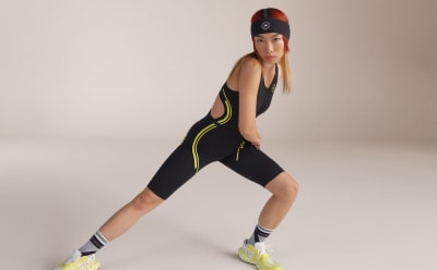 A female model poses wearing the Black and shock Yellow TruePace running one piece. Click on this image to filter the collection by running.