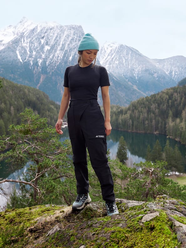 Woman wears black base layer top with black trousers from the Terrex Adventure Wardrobe collection whilst standing on a rock in front of a lake and mountain backdrop.