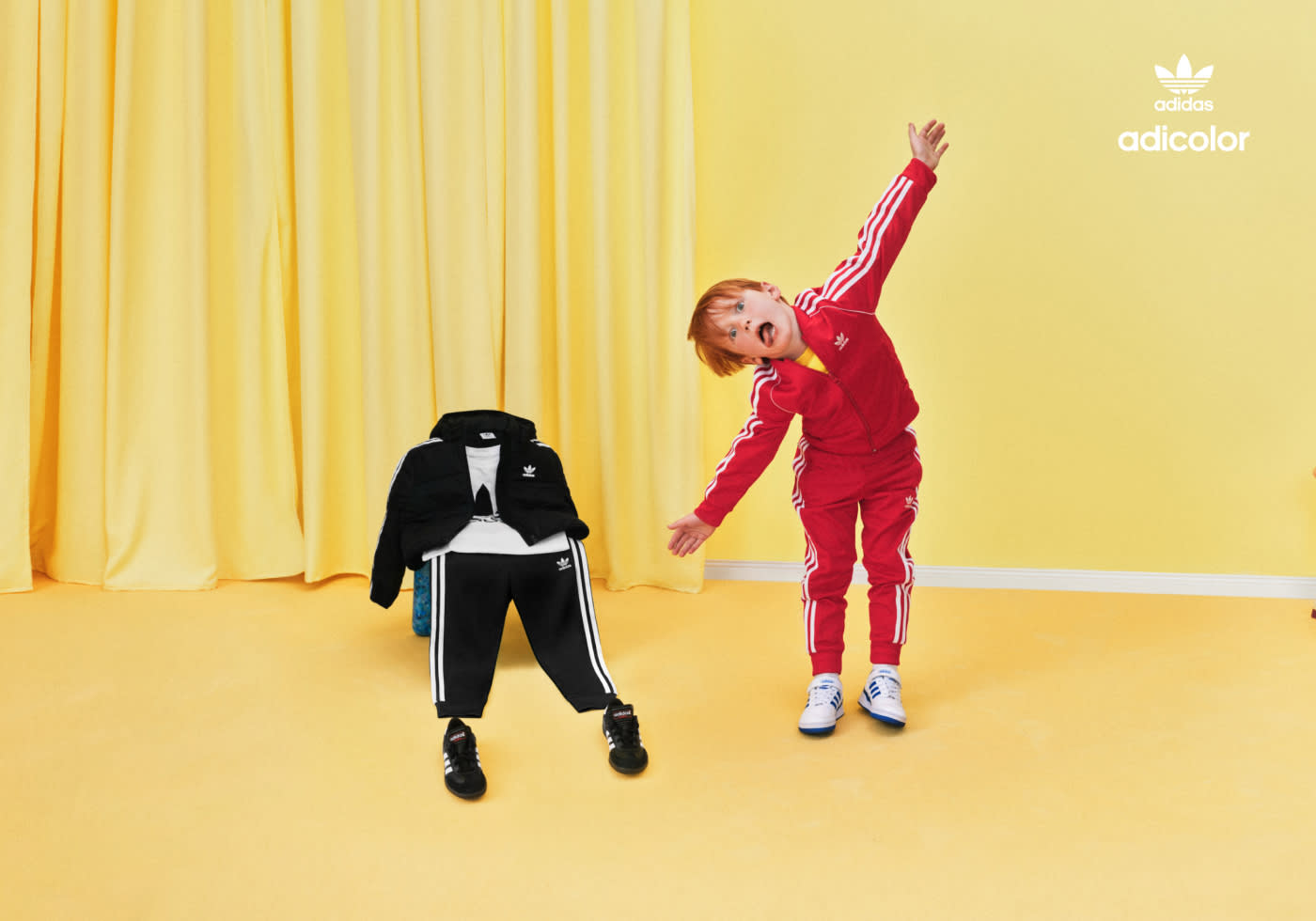 A kid in a yellow room wearing a red adicolor tracksuit, standing next to a chair with a green and black adicolor tracksuit draped over it