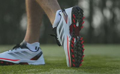 The image shows a close-up view of a person's feet wearing a pair of athletic shoes with red and black accents, standing on a grassy field.
