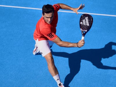 A person in a red shirt is playing tennis on a blue court, with their shadow cast on the ground.