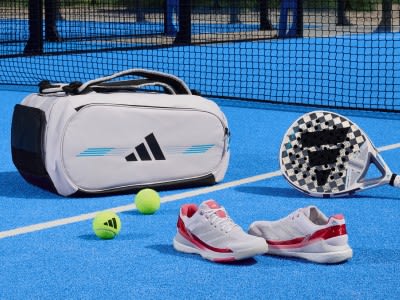 The image shows a sports bag, a tennis racket, and a pair of tennis shoes on a blue tennis court surrounded by a wire fence.