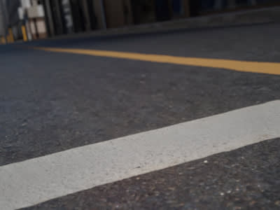 The image shows a close-up view of a road surface, with a white painted line running across the dark asphalt. The background appears to be a blurred urban environment, suggesting this is a street or road in a city or town.