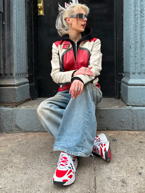 A person with blonde hair wearing a red and white jacket, gray jeans, and red and white sneakers is sitting on steps in front of a building with a metal gate.