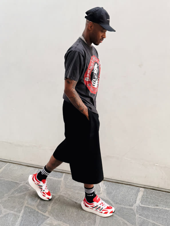 A person wearing a black cap, a graphic t-shirt, and black shorts is standing in a plain, light-colored setting.
