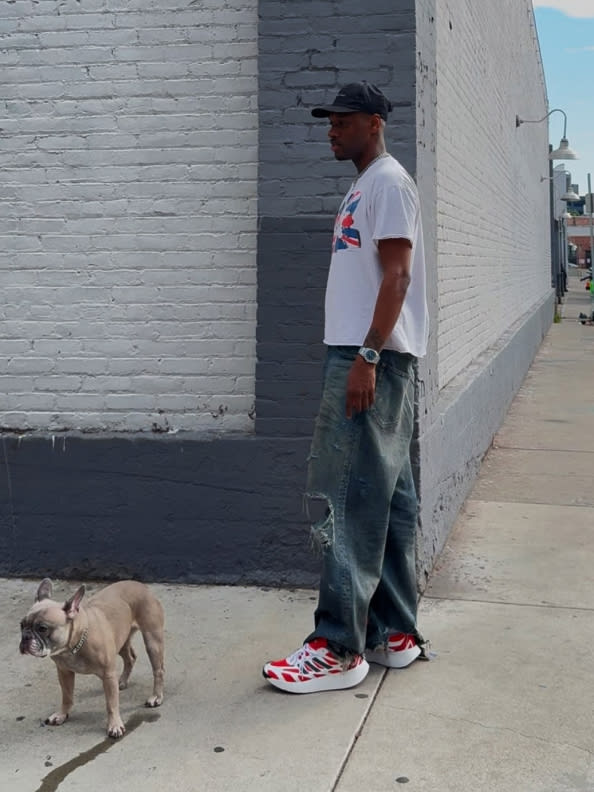 A person wearing a white t-shirt, jeans, and red sneakers stands next to a small dog on a sidewalk in front of a white brick wall.