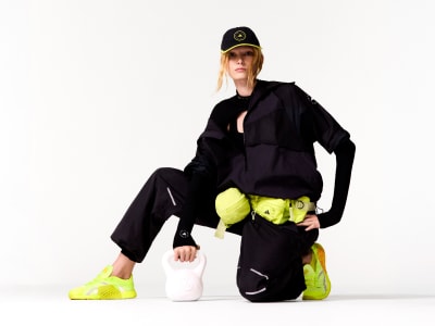 A person wearing a black outfit, including a cap, sits on a white surface against a plain white background, holding yellow objects in their hands.