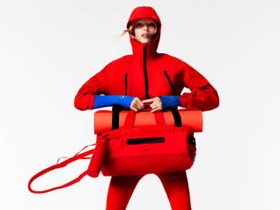 A person wearing a bright red hooded jacket and carrying a large red backpack stands against a plain white background.