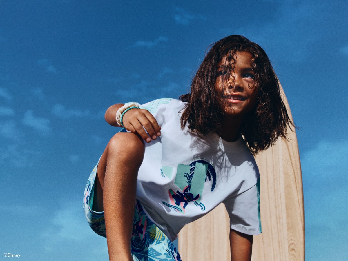 A young person with long dark hair sits on a wooden surface against a clear blue sky, wearing a white t-shirt with a colorful graphic design.