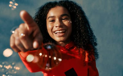A smiling person with curly hair wearing a red jacket stands against a blurred background with sparkling lights.