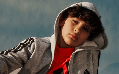 A young person wearing a gray hooded sweatshirt and a red shirt, with a thoughtful expression on their face, set against a blue background.