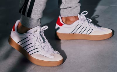 The image shows a pair of white sneakers with red accents and a thick, gum-colored sole, set against a dark background.