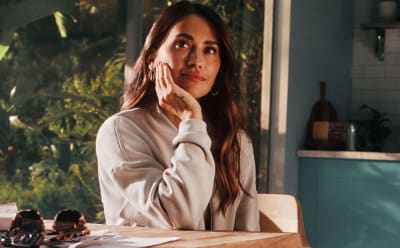 A young woman with long, dark hair sits at a table, her hand covering part of her face, in a cozy indoor setting with a window overlooking greenery in the background.