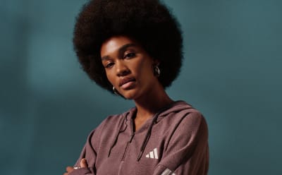A young woman with a large, curly afro hairstyle stands against a dark teal background, wearing a pink hooded sweatshirt.