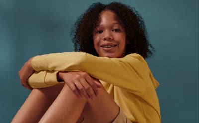 A person with curly dark hair wearing a yellow top sits with their arms wrapped around their knees against a teal background.