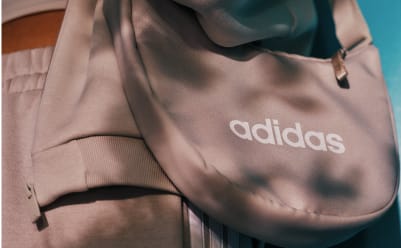 The image shows a close-up view of a pink Adidas backpack, with the Adidas logo prominently displayed on the front. The background appears to be blurred, suggesting the focus is on the backpack itself.
