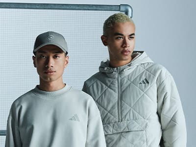Two young men wearing Adidas apparel stand in front of a white wall, one wearing a gray cap and the other a white hooded jacket.
