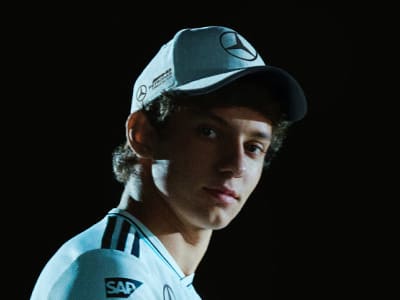 A young man wearing a baseball cap with a logo on it, looking directly at the camera against a dark background.