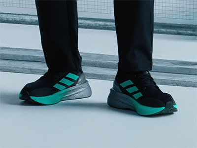 The image shows a pair of black and teal athletic shoes with a distinctive wedge-shaped sole, standing on a snowy surface against a backdrop of a wire mesh fence.