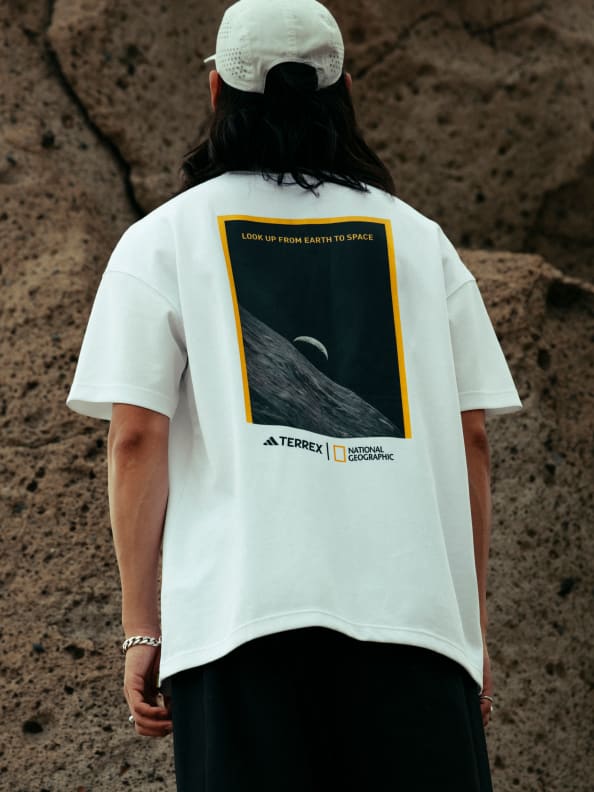 A person wearing a white t-shirt with a graphic design featuring a National Geographic logo and an image of a whale, standing in a rocky outdoor environment.