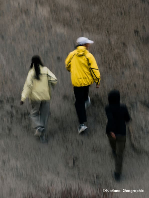 The image depicts several people walking through a rainy field, with one person wearing a bright yellow jacket standing out against the blurred background.