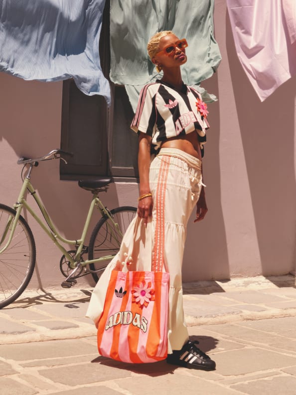 A person wearing a colorful, patterned skirt and a black and white top stands in front of a bicycle and some draped fabric in the background.