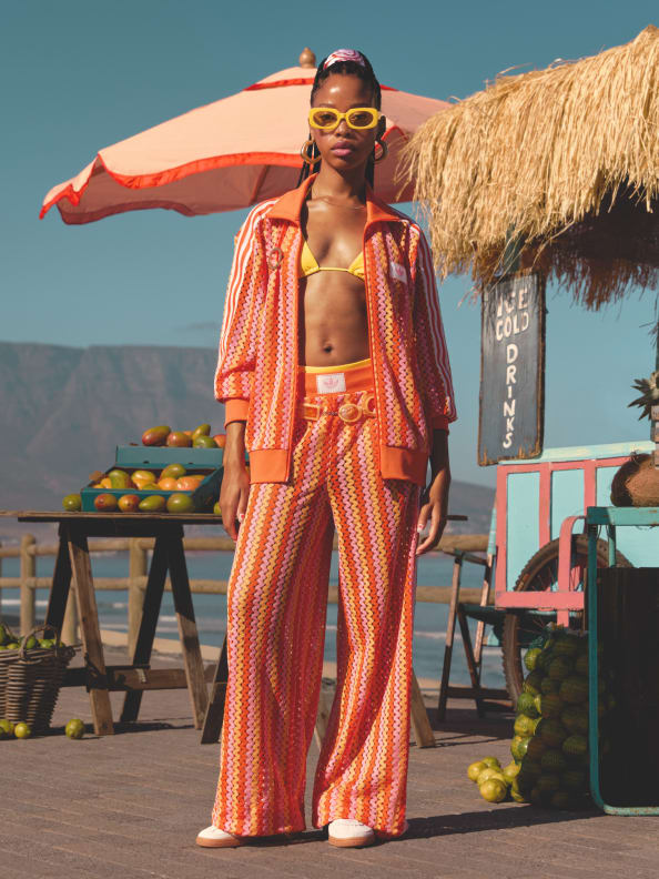 A woman in vibrant, patterned clothing stands in front of a tropical-themed outdoor setting, with a thatched-roof structure and various produce items visible in the background.