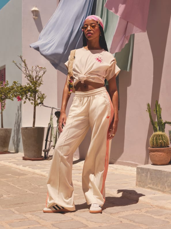 A young woman wearing a white crop top and beige pants stands in a colorful, urban setting with potted plants and a white curtain in the background.