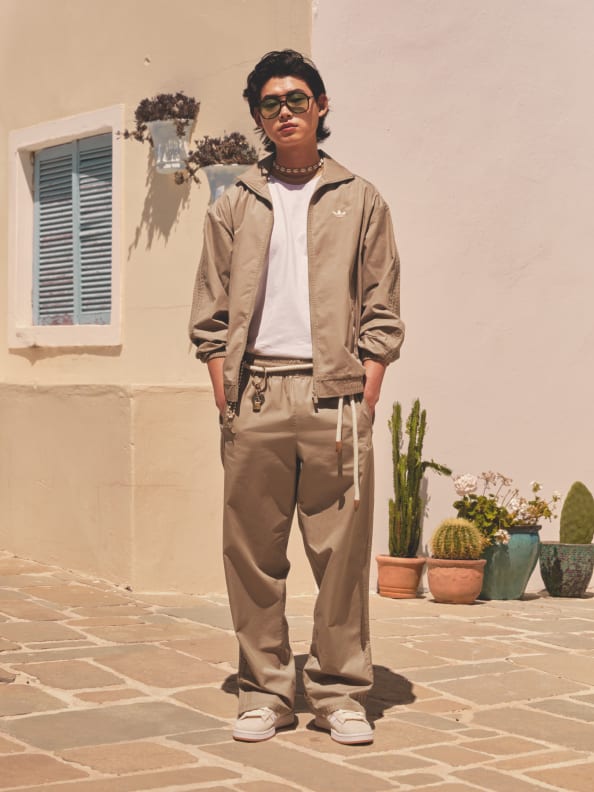 A young man wearing a beige jacket, white shirt, and khaki pants stands in front of a building with a window and potted plants in the background.