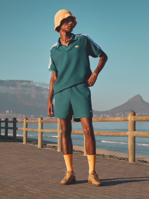 A person wearing a green outfit, including a polo shirt and shorts, stands on a wooden boardwalk overlooking a body of water with mountains in the background.