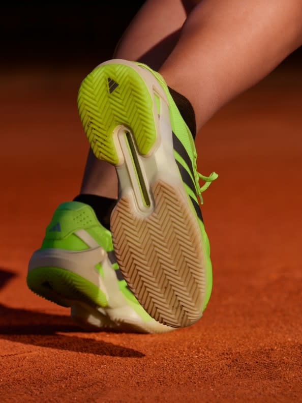 A pair of bright green and white athletic shoes with a distinctive sole pattern are visible on a reddish-brown surface, likely a tennis court.