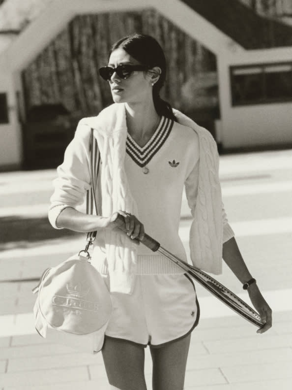 A person wearing a white athletic outfit and sunglasses, holding a tennis racket, stands on a tennis court with a building visible in the background.