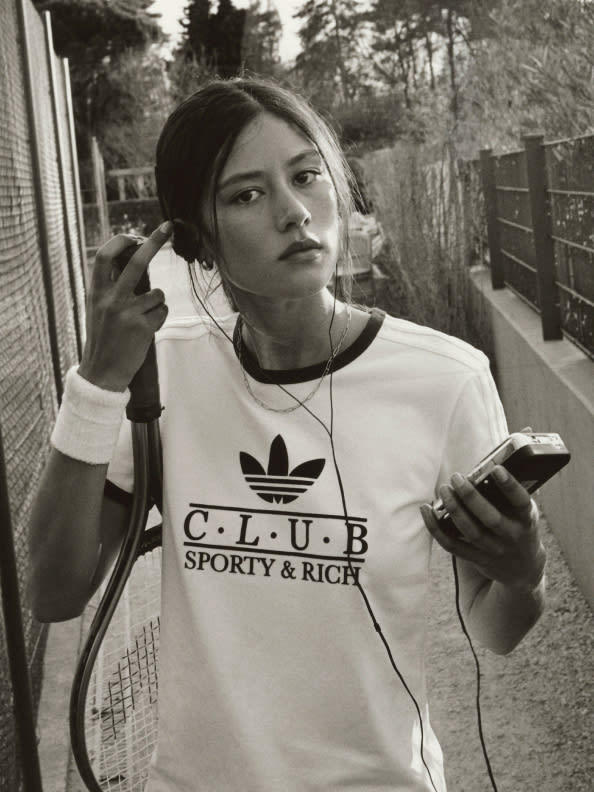 A young woman wearing a white t-shirt with a logo stands in front of a wooden fence, holding a mobile device and looking thoughtful.