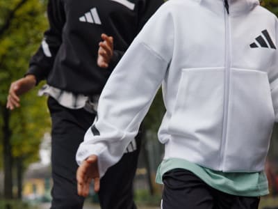 The image shows a person wearing a white and black Adidas jacket, standing in a grassy outdoor setting.