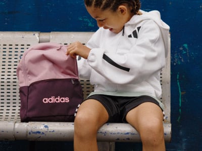 A young person in a white athletic jacket and shorts is sitting on a bench, with a pink and burgundy Adidas backpack next to them.