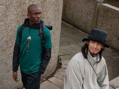 Two individuals, a man wearing a green sweater and a woman wearing a gray coat and hat, stand in an urban setting with a concrete wall in the background.