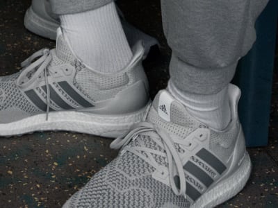 The image shows a pair of white and gray Adidas sneakers with laces and the Adidas logo visible, set against a dark background.