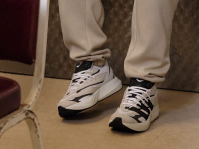The image shows a pair of white and black sneakers on a person's feet, with the person wearing beige pants in a room with a patterned carpet.