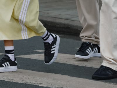 The image shows a pair of black and white sneakers with distinctive stripes, worn by two individuals standing on a concrete surface.